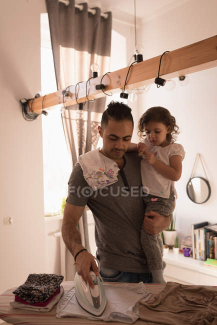 Man ironing cloth while carrying daughter at home. — Stock Photo