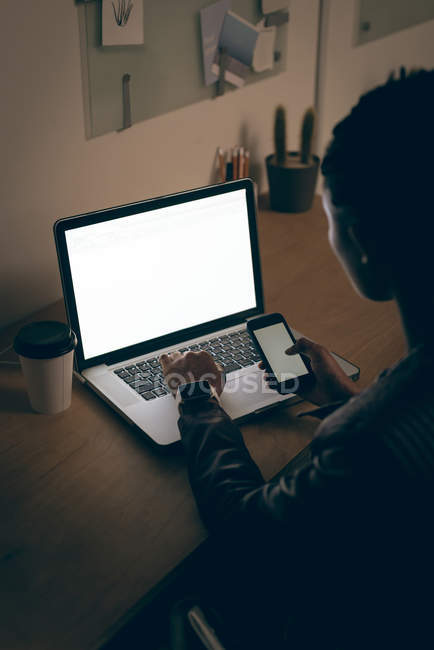 Weibliche Führungskraft benutzt Handy, während sie am Schreibtisch im Büro am Laptop arbeitet — Stockfoto
