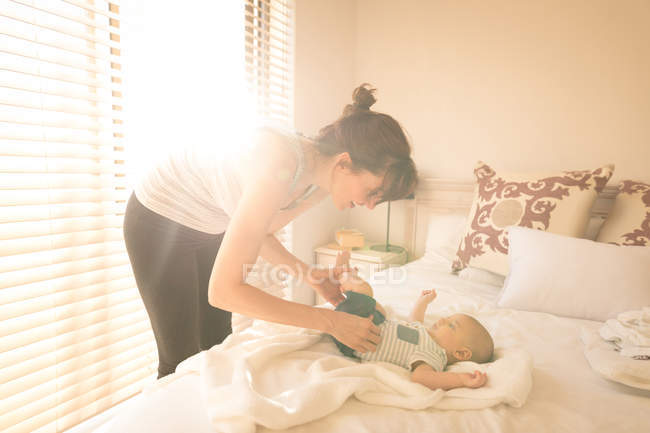 Lindo bebé vistiéndose por su madre en la cama en el dormitorio en casa - foto de stock