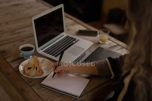 Reife Frau sitzt auf Stuhl und schreibt zu Hause auf Notizbuch — Stockfoto