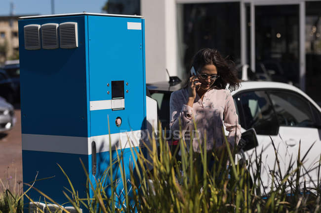 Mulher falando no telefone celular enquanto carrega carro elétrico na estação de carregamento — Fotografia de Stock