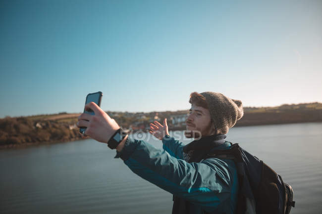 Caminhante masculino levando selfie com telefone celular perto do lago no campo — Fotografia de Stock
