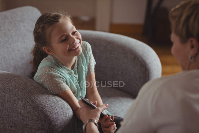 Età elementare ragazza in possesso di smartphone mentre interagisce con la madre in soggiorno . — Foto stock