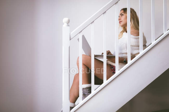 Mujer reflexiva usando el ordenador portátil mientras está sentado en la escalera en casa - foto de stock