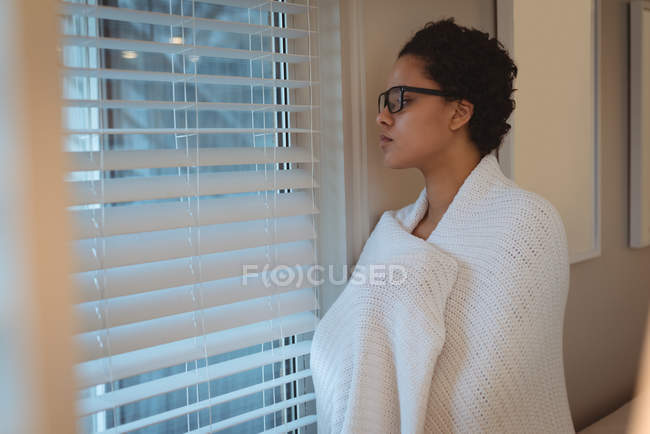 Jeune femme enveloppée dans une couverture regardant par la fenêtre à la maison — Photo de stock