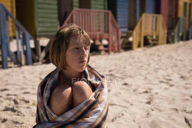 Thoughtful teenage girl wrapped in blanket sitting on beach — Stock Photo