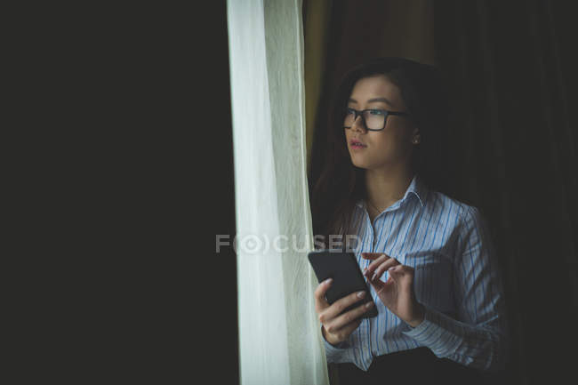 Businesswoman looking through window while using mobile phone at hotel room — Stock Photo