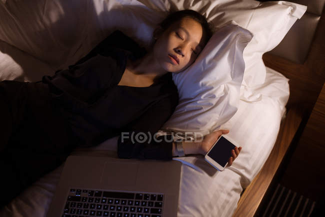 Уставшая женщина спит с ноутбуком и мобильным телефоном на кровати в отеле — стоковое фото