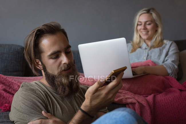 Homem falando no telefone celular enquanto mulher usando laptop na sala de estar em casa — Fotografia de Stock