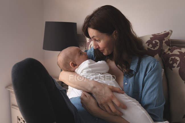 Joyeux jeune mère assise sur le lit regardant bébé à la maison — Photo de stock