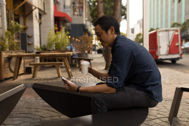 Sonriente hombre de negocios asiático utilizando el teléfono móvil en la cafetería del pavimento - foto de stock