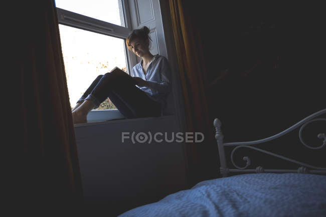 Donna che legge libro sul davanzale della finestra in camera da letto a casa — Foto stock