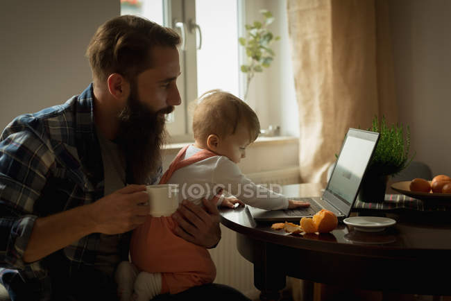Vater trinkt Kaffee, während die Sonne zu Hause am Laptop spielt — Stockfoto