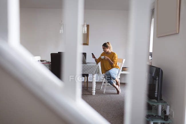 Mujer usando el teléfono móvil mientras toma café en casa - foto de stock