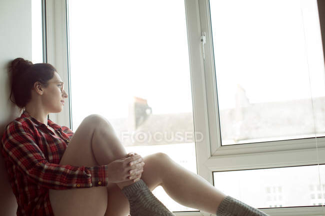 Donna pensierosa in occhiali seduta sul davanzale della finestra a casa . — Foto stock