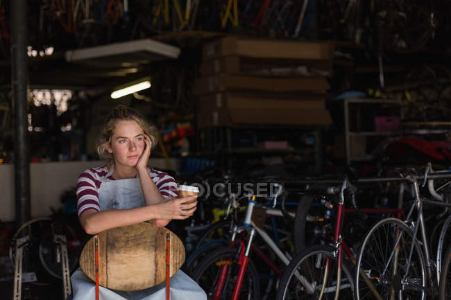Thoughtful female mechanic having coffee in workshop — Stock Photo