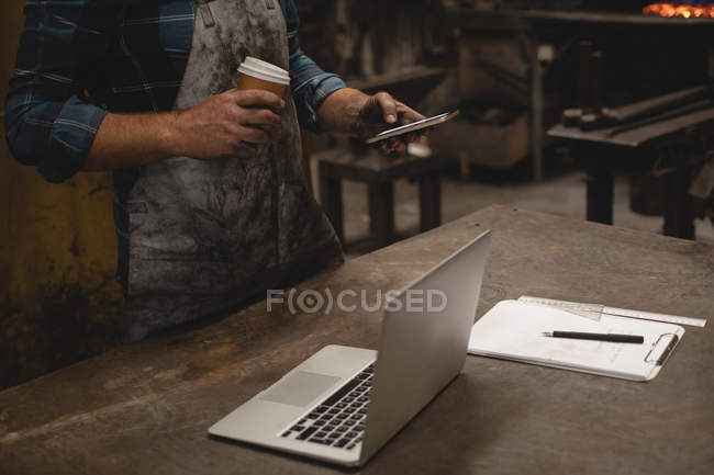 Mittlerer Teil des Schmieds beim Kaffeetrinken mit Handy in Werkstatt — Stockfoto
