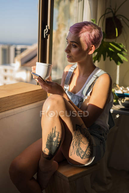 Elegante donna con capelli rosa in possesso di caffè e telefono cellulare alla luce del sole a casa . — Foto stock