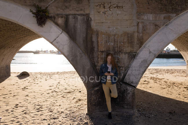 Жінка, використовуючи мобільний телефон, спираючись на міст у сонячному світлі — стокове фото