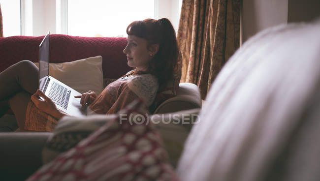 Mujer usando el ordenador portátil mientras se relaja en el sofá en la sala de estar en casa - foto de stock
