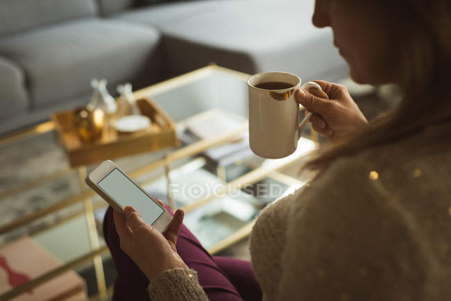 Mulher usando telefone celular enquanto toma café em casa — Fotografia de Stock