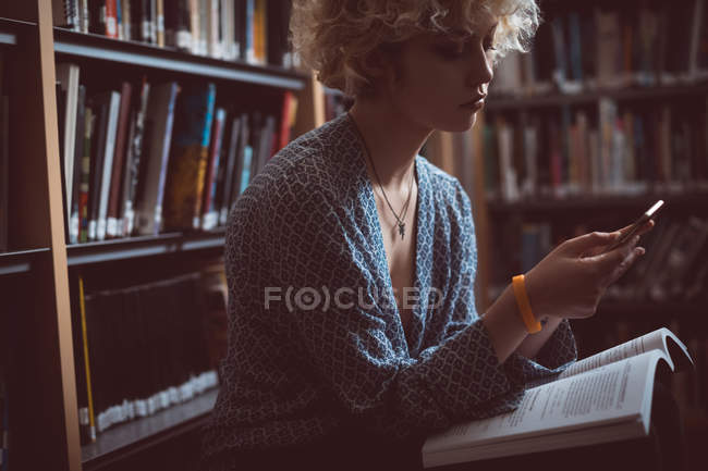 Mulher usando telefone celular enquanto lê um livro na biblioteca — Fotografia de Stock
