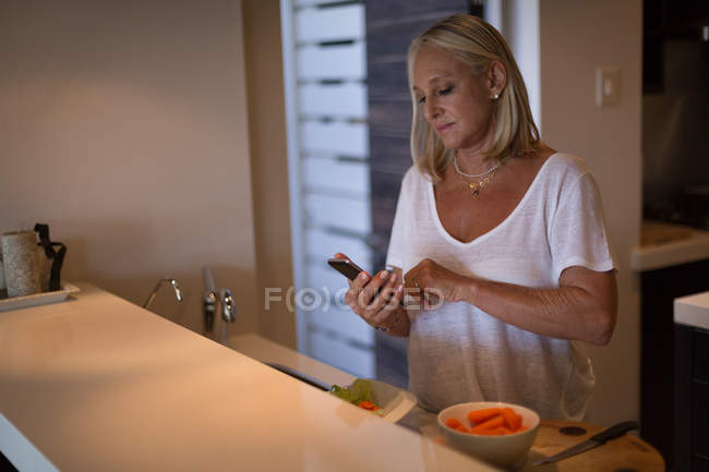 Femme blonde mature utilisant le téléphone mobile dans la cuisine à la maison . — Photo de stock