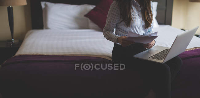 Mujer de negocios leyendo documentos mientras trabaja en el ordenador portátil en la habitación de hotel - foto de stock