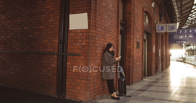 Mujer usando teléfono móvil contra pared de ladrillo en la estación de tren - foto de stock