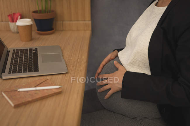 Беременная деловая женщина, прикасающаяся к своему животу за столом в офисе — стоковое фото