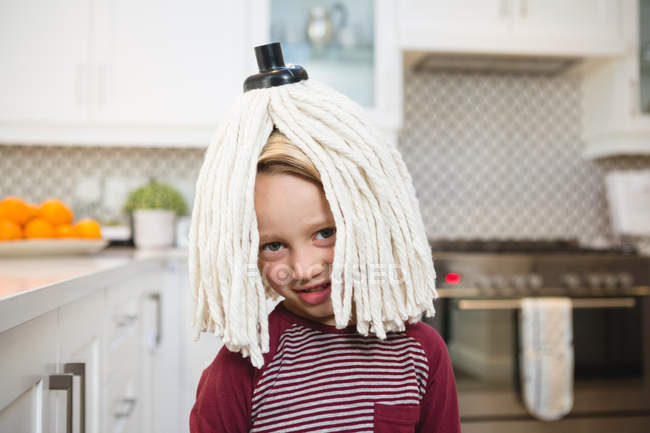 Niño feliz con fregona en la cabeza en la cocina en casa - foto de stock