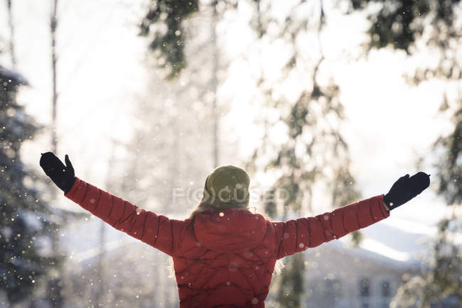 Visão traseira da mulher feliz em pé com os braços estendidos durante a queda de neve — Fotografia de Stock