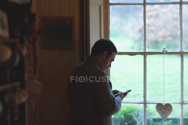 Uomo che utilizza il telefono cellulare vicino alla finestra a casa — Foto stock
