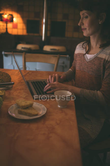 Mujer usando el ordenador portátil mientras desayuna en casa - foto de stock