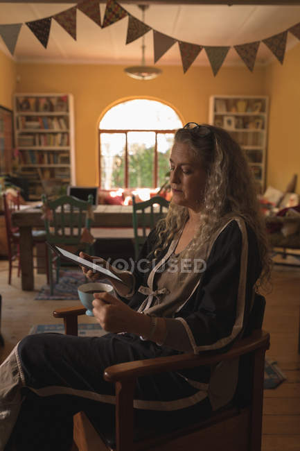 Mulher madura usando tablet digital enquanto toma café em casa — Fotografia de Stock