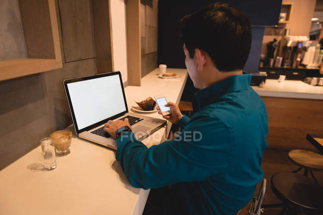Чоловік використовує ноутбук і мобільний телефон, сидячи в кав'ярні — стокове фото