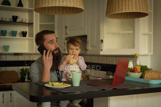 Father holding baby while talking on mobile phone at home — Stock Photo