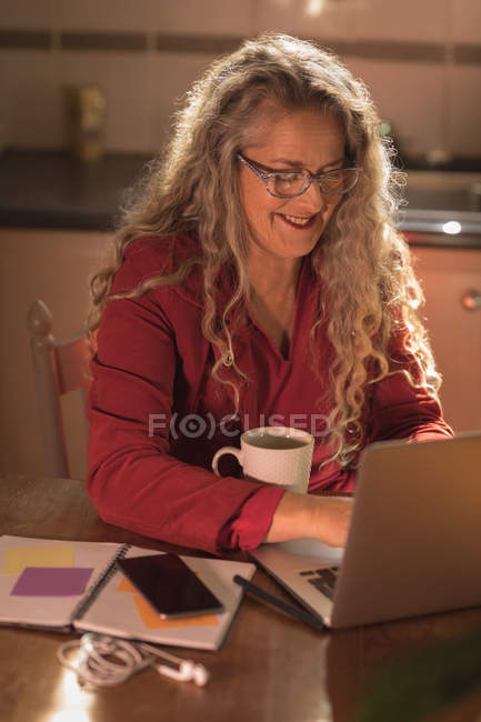 Усміхнена зріла жінка сидить на стільці і використовує свій ноутбук вдома — стокове фото