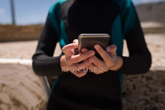 Средняя секция девушки в мокром костюме с помощью мобильного телефона на пляже — стоковое фото