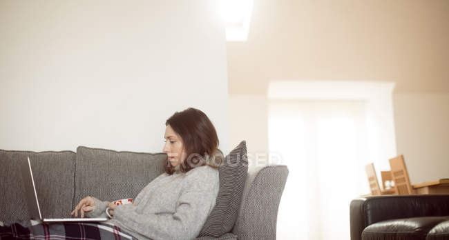 Mulher segurando xícara de café ao usar laptop no sofá na sala de estar em casa . — Fotografia de Stock