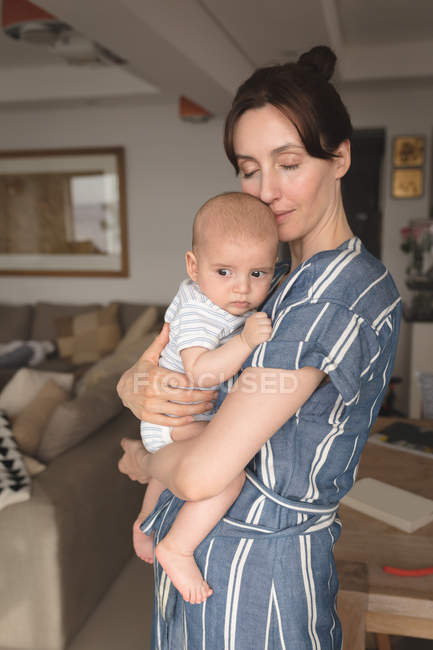 Mamá con los ojos cerrados sosteniendo a su bebé en la sala de estar en casa - foto de stock