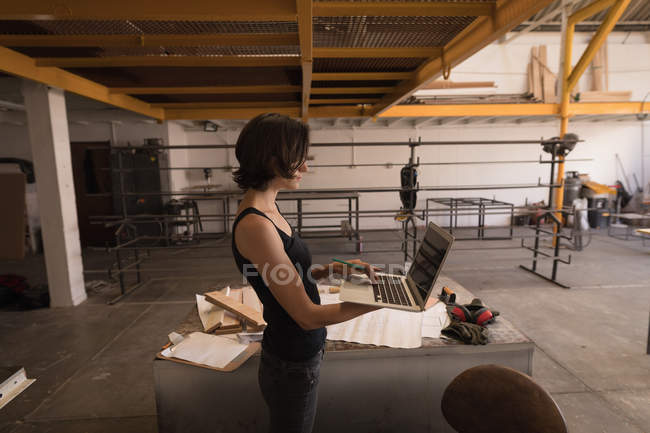 Junge Handwerkerin nutzt Laptop in Werkstatt. — Stockfoto