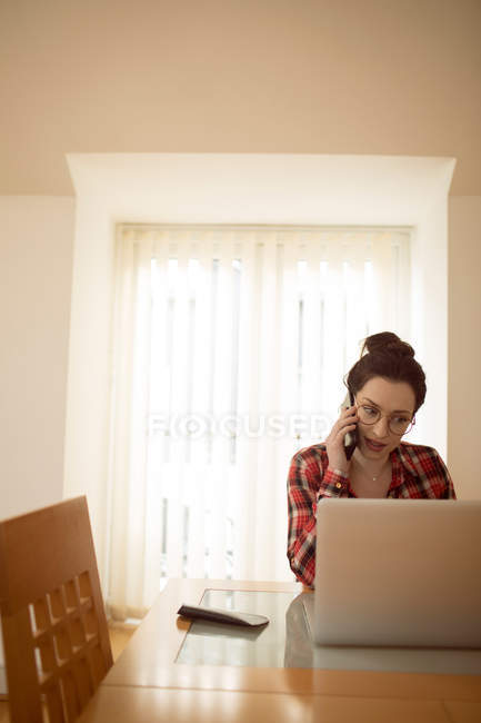 Mulher falando no celular enquanto usa laptop em casa. — Fotografia de Stock