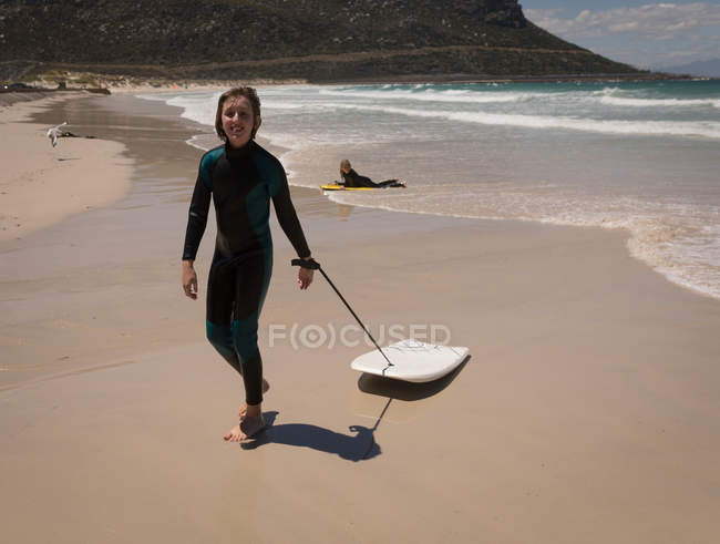 Portrait of teenage girl in wet suit walking with surfboard on beach — Stock Photo