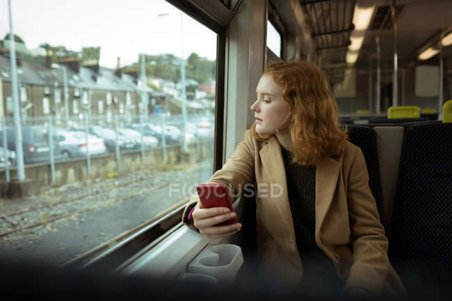 Mujer joven pelirroja mirando hacia otro lado mientras usa su móvil en tren - foto de stock