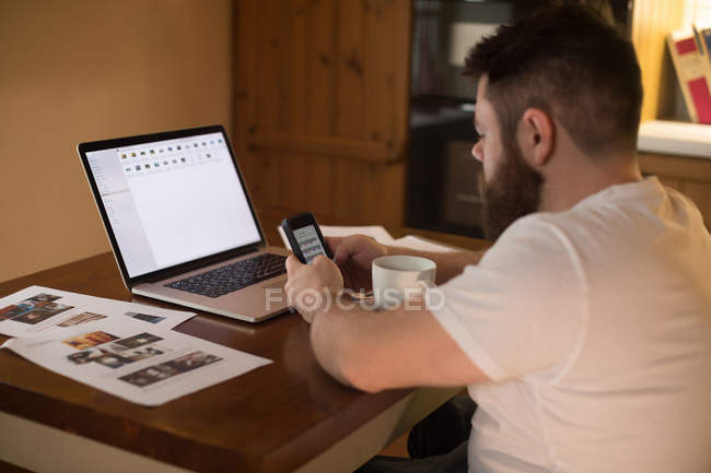 Людина з обмеженими можливостями використовує мобільний телефон під час роботи на ноутбуці вдома — стокове фото