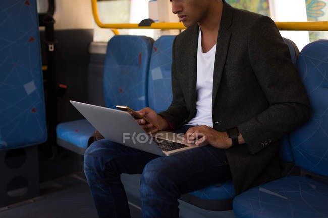 Sección media del hombre de negocios que usa el teléfono móvil y el portátil mientras viaja en autobús - foto de stock