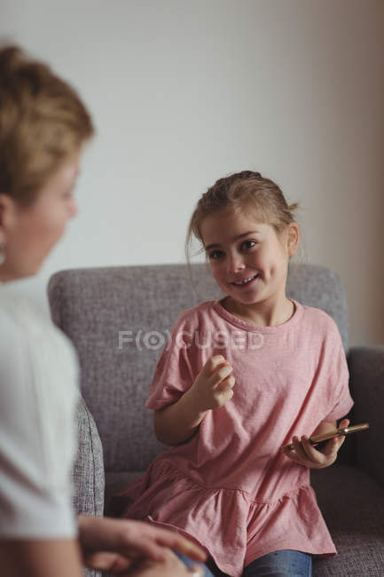Mädchen im Grundschulalter hält Smartphone in der Hand, während sie mit Mutter im Wohnzimmer interagiert. — Stockfoto