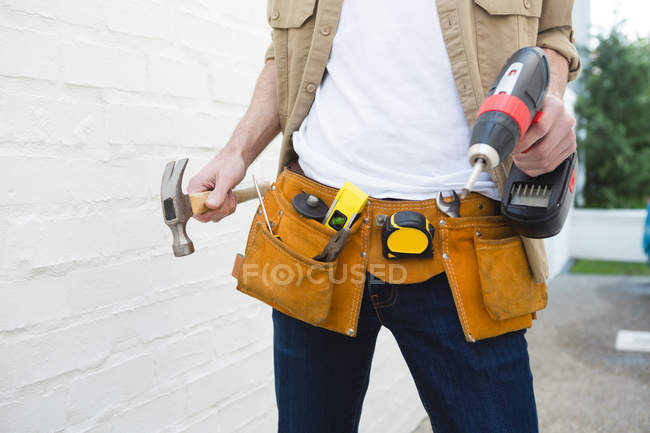 Seção média de carpinteiro masculino com cinto de ferramentas segurando martelo e máquina de broca — Fotografia de Stock