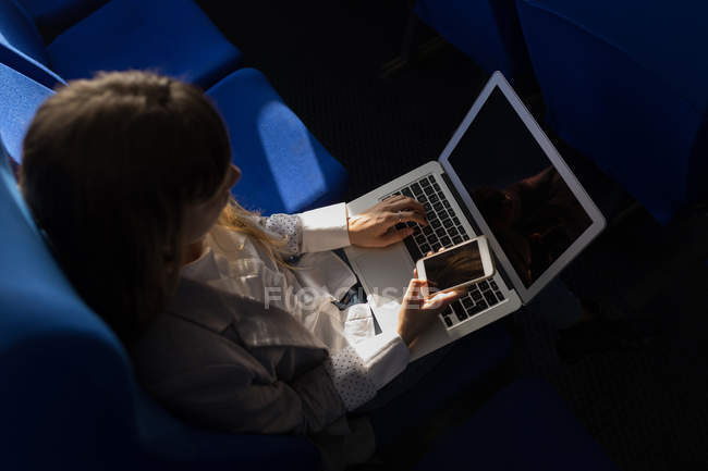 Hochwinkelaufnahme einer Frau, die im Kreuzfahrtschiff ihr Handy kontrolliert, während sie Laptop benutzt — Stockfoto